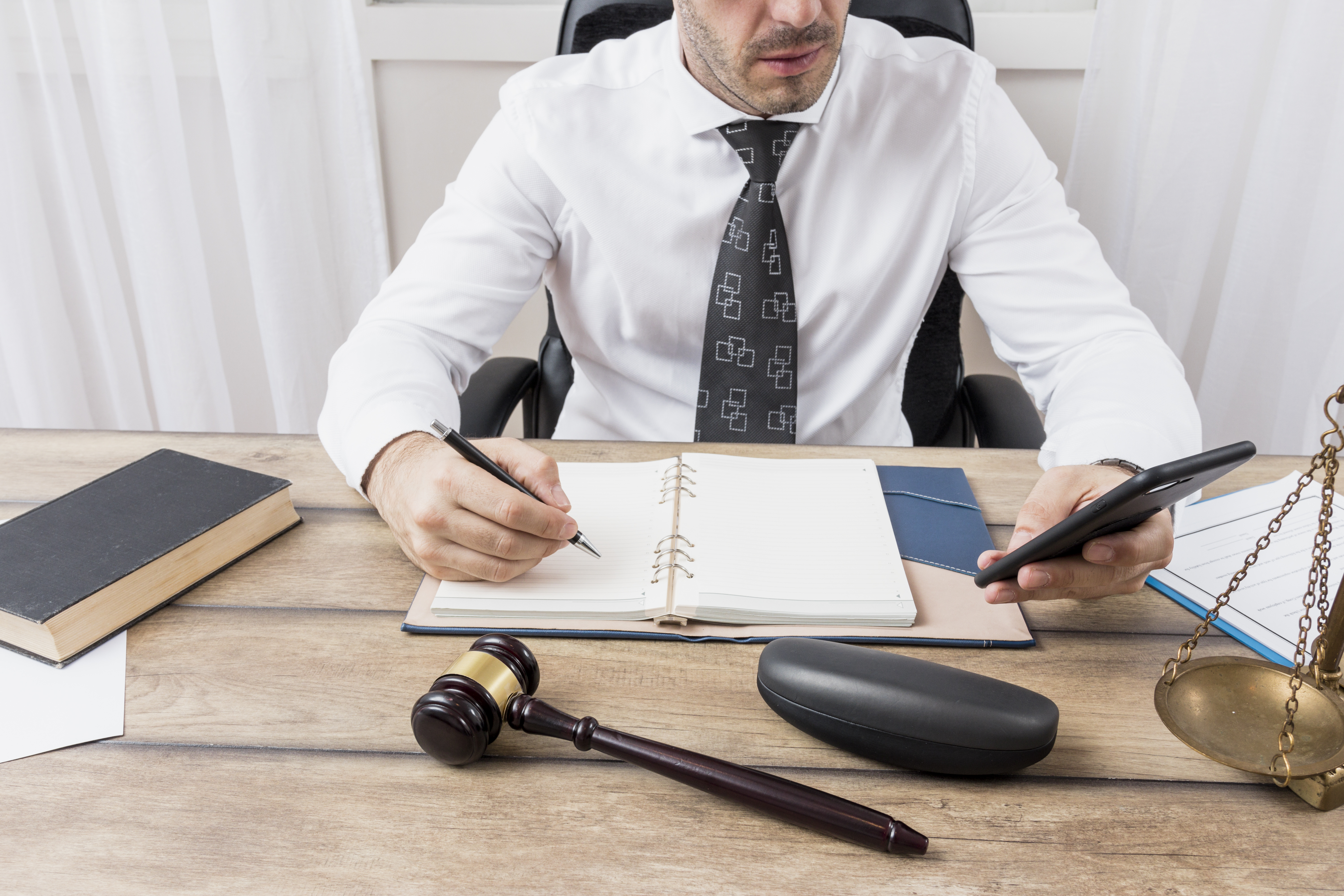 A lawyer sitting at his desk, checking his smartphone and writing on a notebook.
